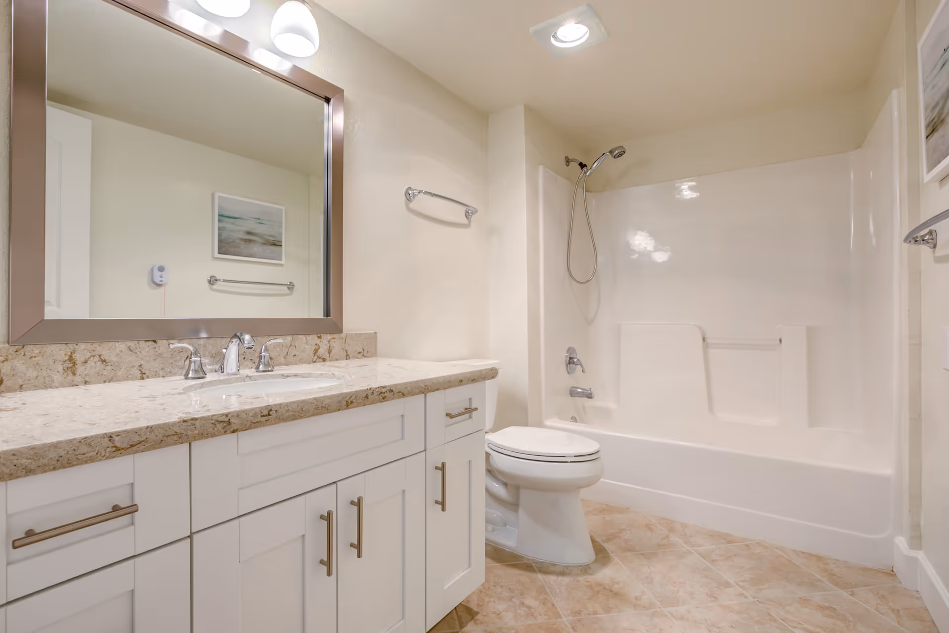 A clean and modern bathroom featuring a white vanity with a beige marble countertop, a large rectangular mirror with a silver frame, a toilet, and a white bathtub with a showerhead. The floor is tiled with beige tiles, and the walls are painted light beige. There is a towel rack on the wall and a framed picture reflected in the mirror.