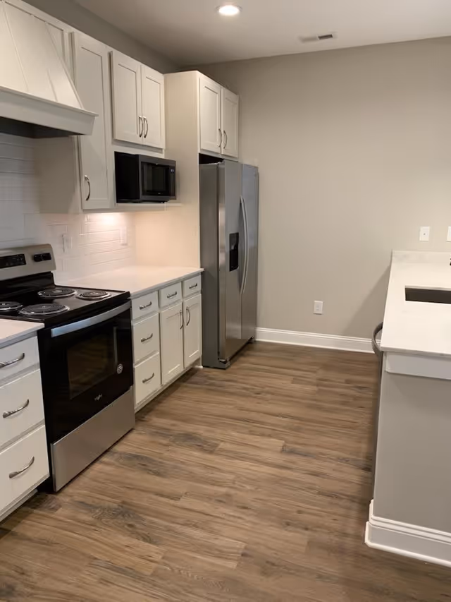Modern kitchen with white cabinets, stainless steel refrigerator and oven, microwave, white countertops, and wood flooring.