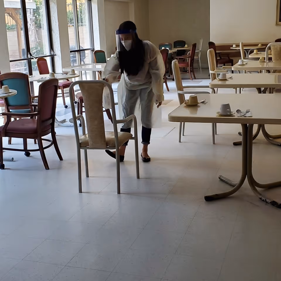 A person wearing protective gear including a face shield, mask, and gown is arranging a chair in a dining area with multiple tables and chairs. The tables have cups and utensils set on them, and large windows allow natural light into the room.