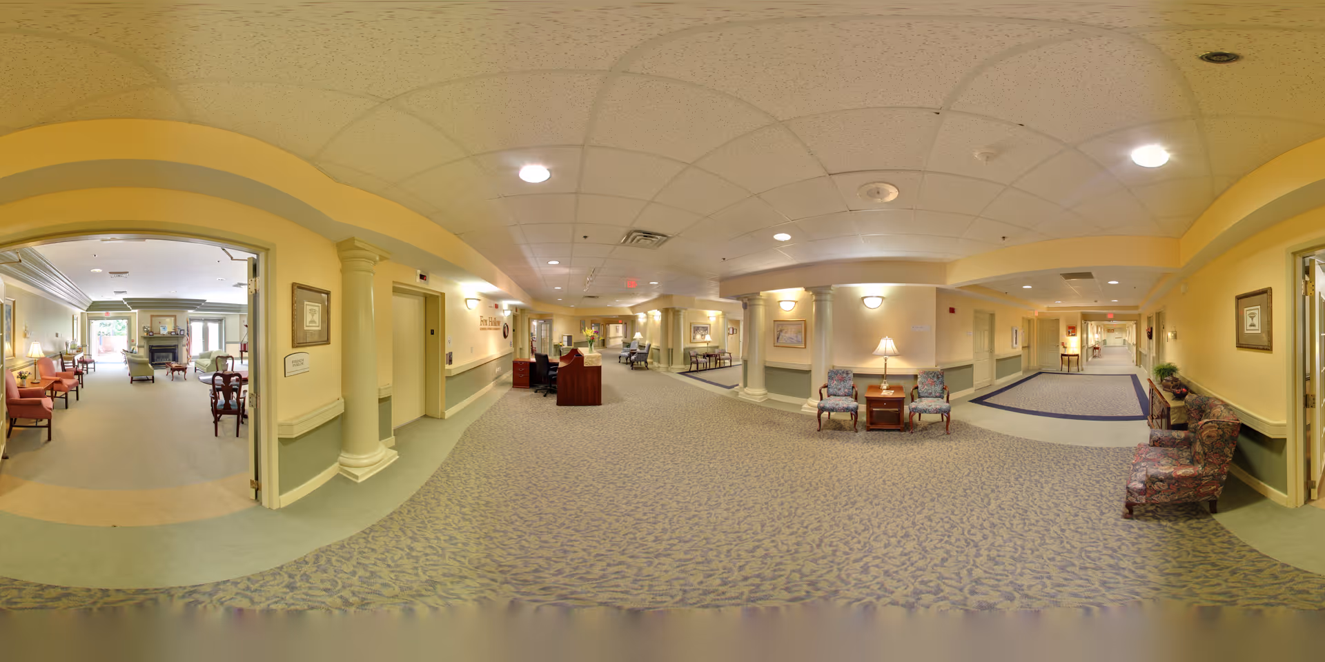 Wide panoramic view of a senior living facility interior hallway with soft yellow walls, patterned carpet, and ceiling lights. The hallway features seating areas with armchairs and side tables with lamps. On the left, there is an open doorway leading to a common living room area with more chairs and a fireplace. The space is well-lit and designed for comfort and accessibility.