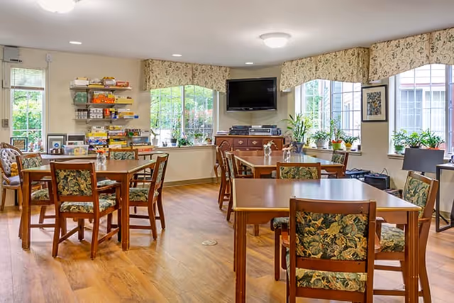 A bright and cozy common area with multiple wooden tables and chairs featuring floral upholstery. Large windows with floral valances allow natural light to fill the room. A flat-screen TV is mounted on the wall above a wooden cabinet, and shelves with board games are visible in the corner. Several potted plants are placed on the windowsills, adding a touch of greenery.