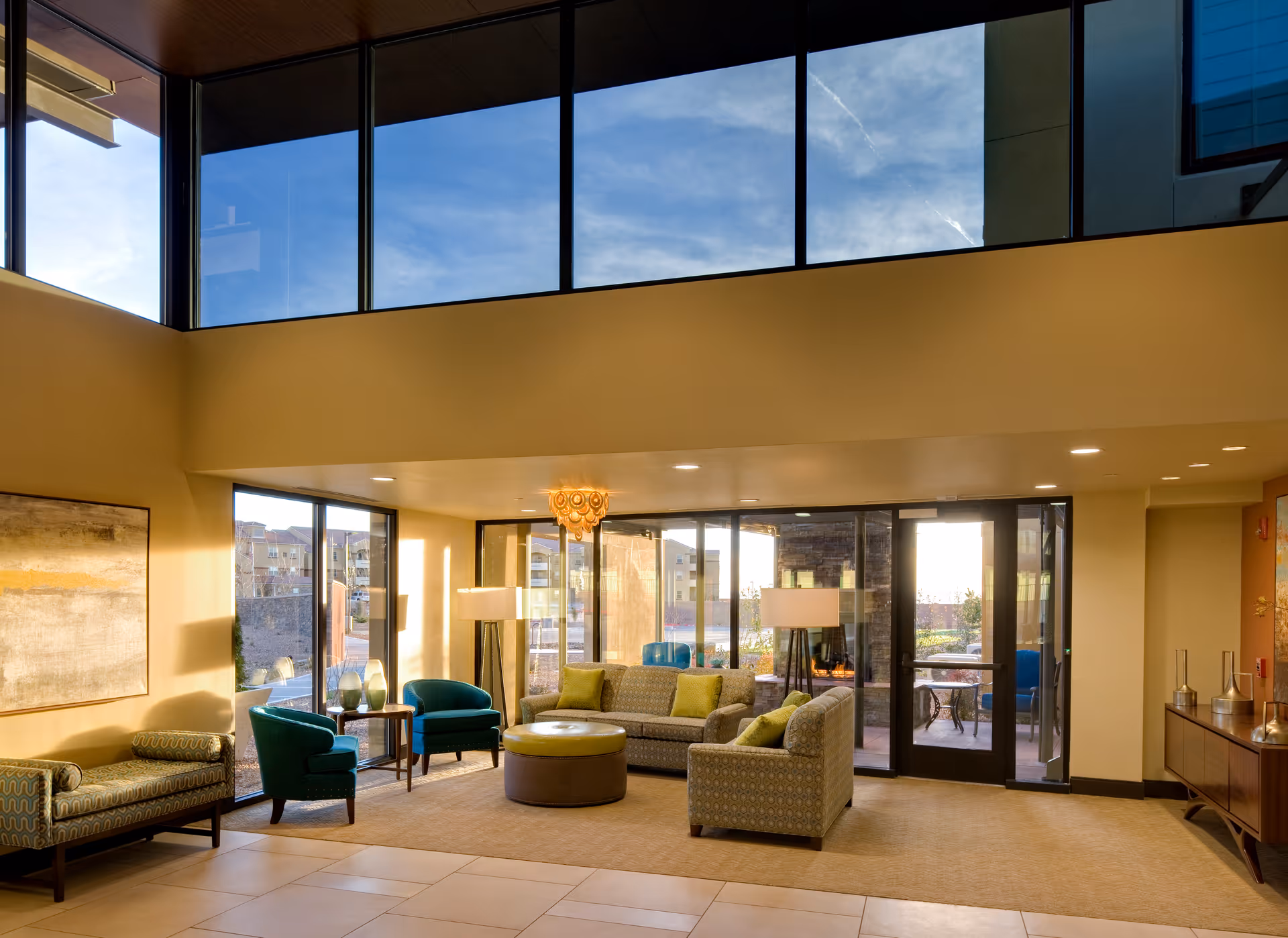 A spacious and well-lit living room area with large floor-to-ceiling windows showing an outdoor patio. The room features a patterned sofa with green cushions, two teal armchairs, a round ottoman, a bench with patterned upholstery, two floor lamps, a wooden sideboard, and a modern chandelier. The walls are painted a warm beige color, and there is a large abstract painting on one wall.