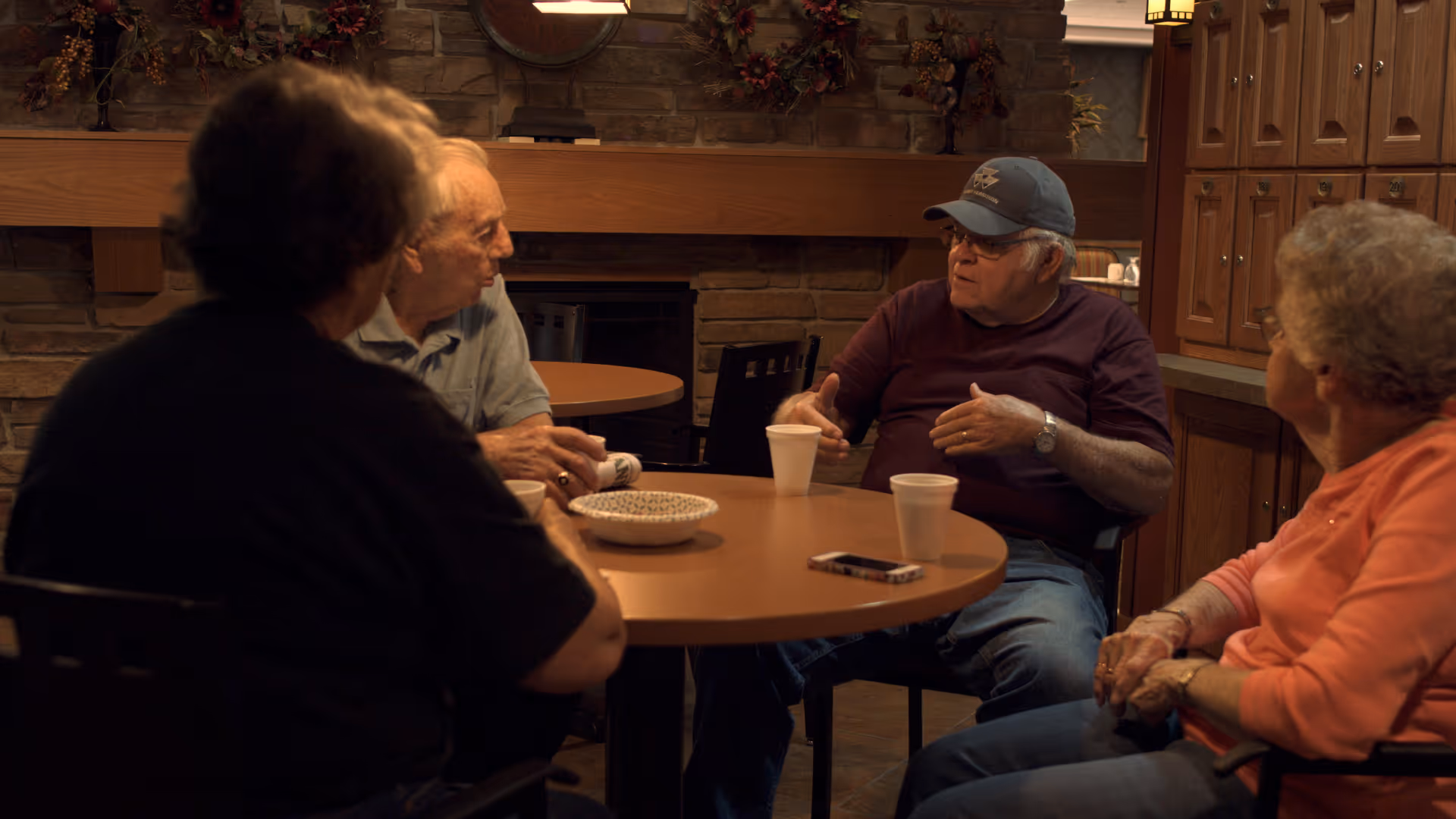 Four elderly people sit around a round table chatting in a cozy common room with a fireplace.