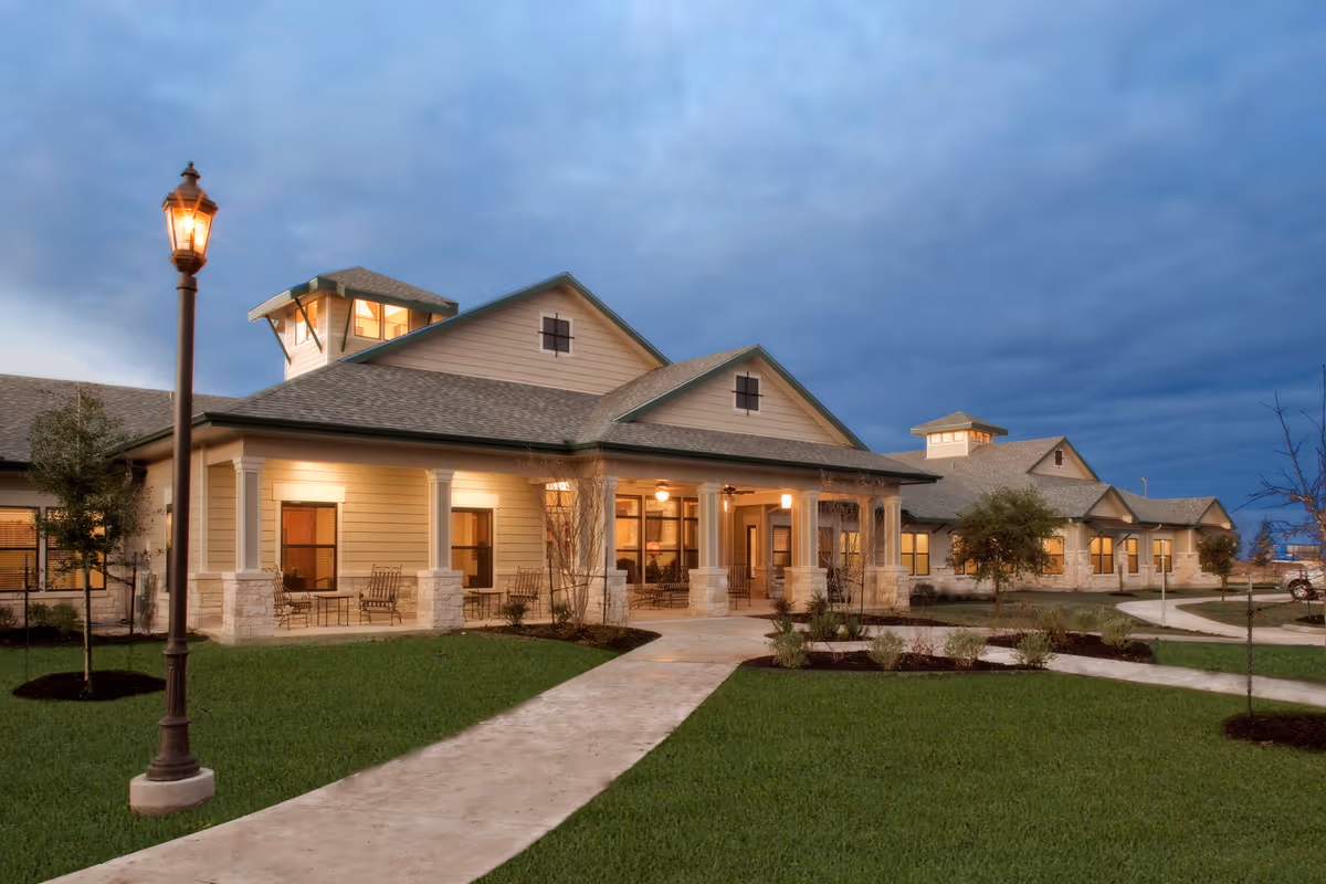 Exterior view of a single-story assisted living facility building at dusk with warm lights glowing from inside. The building has a beige siding with stone accents and a covered porch with seating. A lit streetlamp stands on a concrete pathway leading to the entrance, surrounded by green grass and small landscaped trees and bushes.