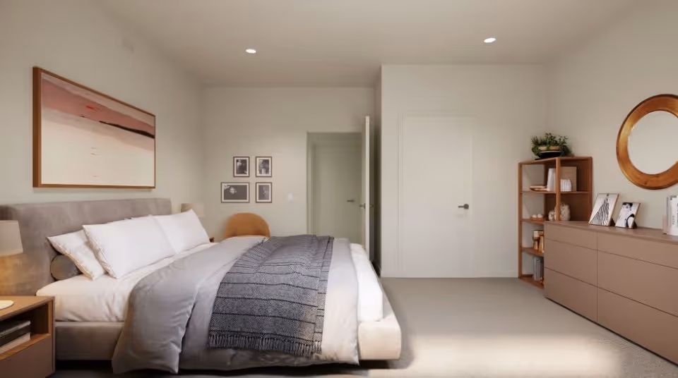 A modern bedroom with a large bed featuring white pillows and gray bedding. There is a wooden nightstand with a lamp on the left side of the bed. On the wall above the bed is a large abstract painting. To the right, there is a wooden dresser with a round mirror above it and a small shelving unit with decorative items and plants. The room has beige walls and carpeted flooring, with two closed white doors visible in the background.
