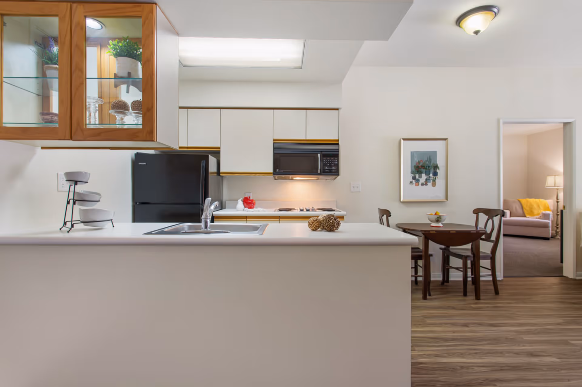 A modern kitchen with a white countertop island featuring a sink and faucet. Behind the island, there is a black refrigerator, white cabinets, a microwave, and a stove with a red kettle. To the right, a small wooden dining table with two chairs is set against a white wall with a framed plant artwork. Through an open doorway, a living room with a sofa, a yellow throw blanket, and a floor lamp is visible.