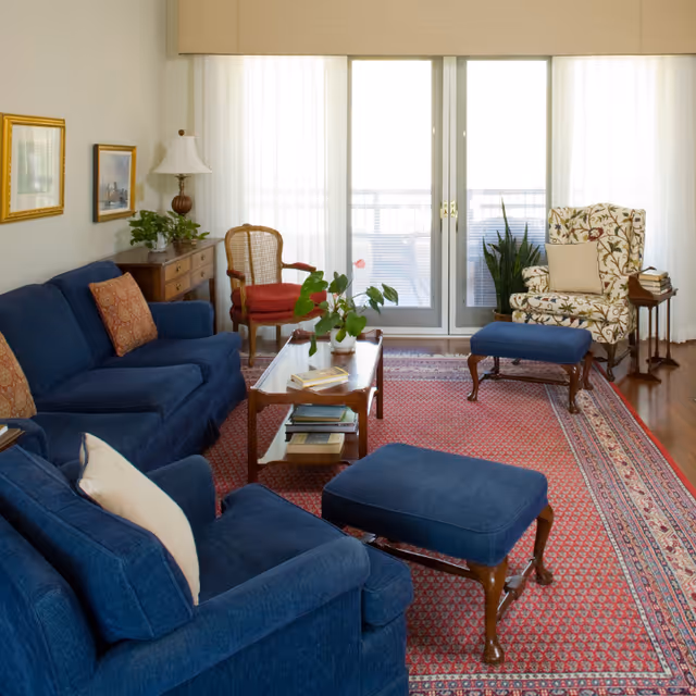 A cozy living room with blue upholstered sofas and ottomans, a wooden coffee table with books and a plant, a floral armchair with a beige pillow, a wooden side table with books, a red cushioned chair, and large glass doors with sheer white curtains letting in natural light.