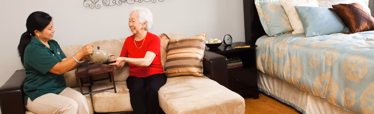 A caregiver in a green shirt is serving tea to an elderly woman wearing a red shirt, both sitting on a beige sectional sofa in a cozy room with a bed and nightstand visible in the background.