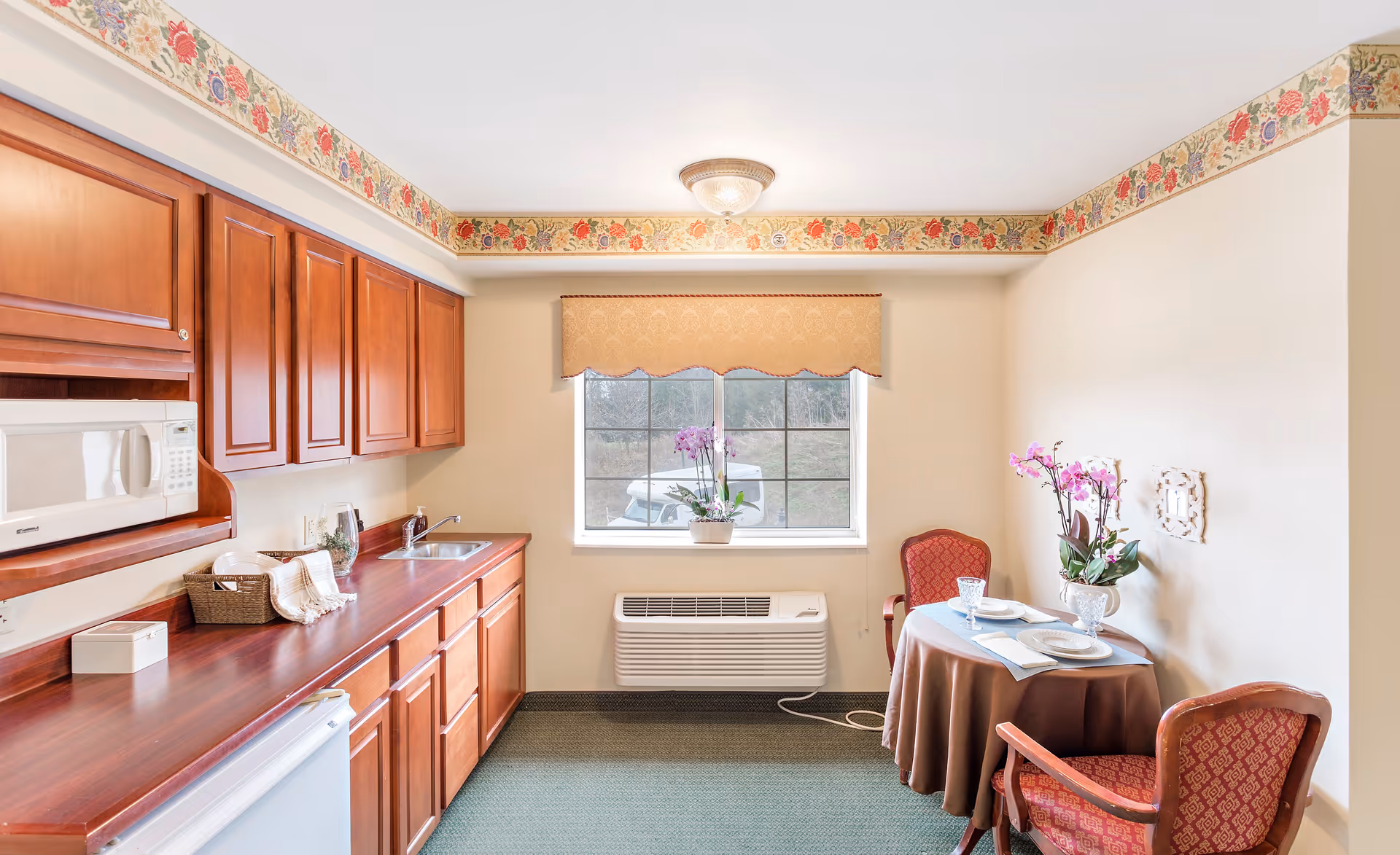 A small kitchen and dining area in a retirement facility with wooden cabinets, a microwave, a sink, and a countertop on the left side. A window with a beige valance is centered on the back wall, with a potted orchid on the windowsill. Below the window is a wall-mounted air conditioning unit. On the right side, there is a round table covered with a brown tablecloth, set with two plates, glasses, and napkins, accompanied by two red upholstered chairs. The room has floral wallpaper border near the ceiling and light-colored walls.
