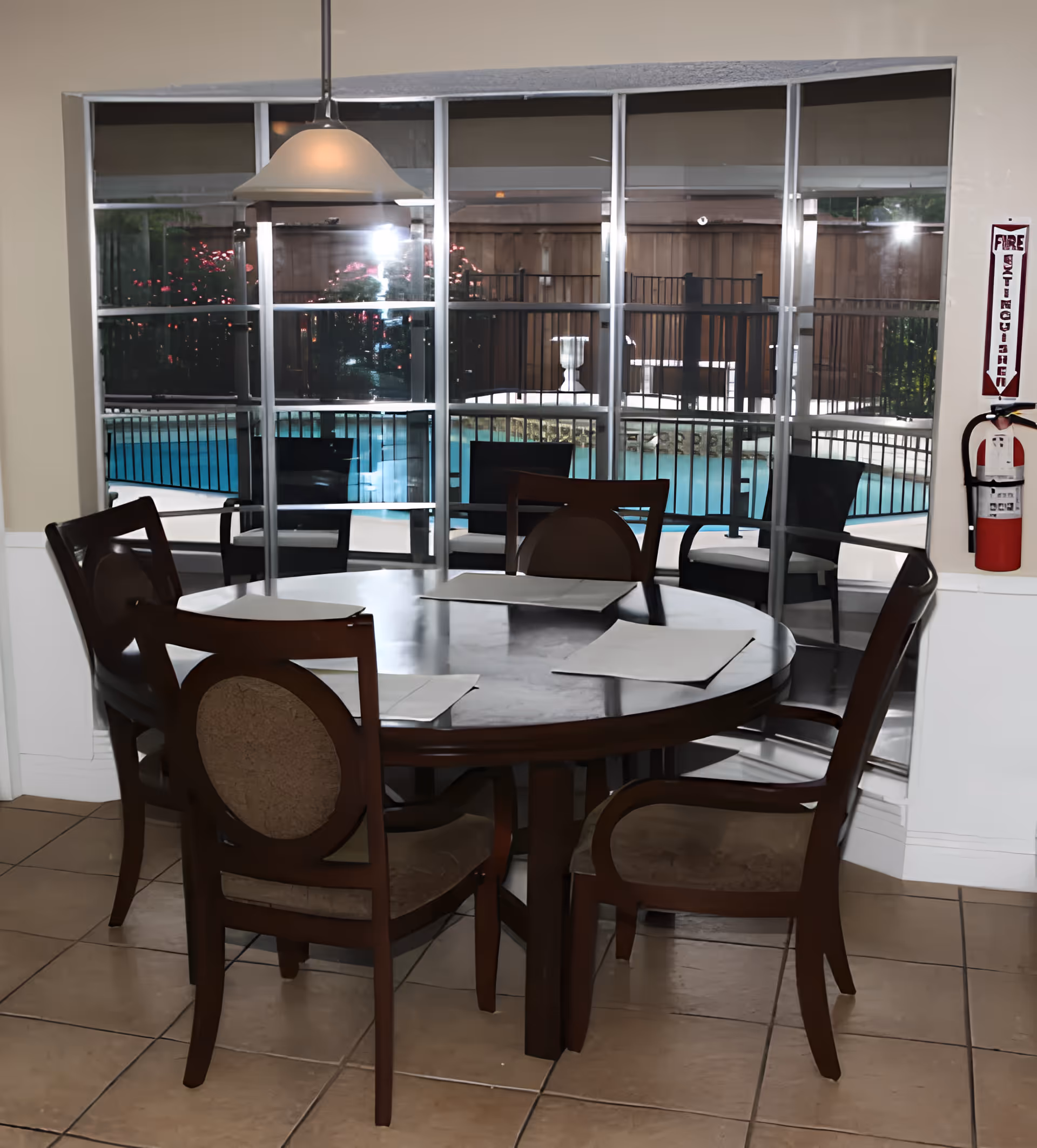 A round wooden dining table with six chairs around it, set with placemats, located indoors near a large window. Outside the window, there is a fenced swimming pool and some greenery. A hanging light fixture is above the table, and a fire extinguisher is mounted on the wall to the right.