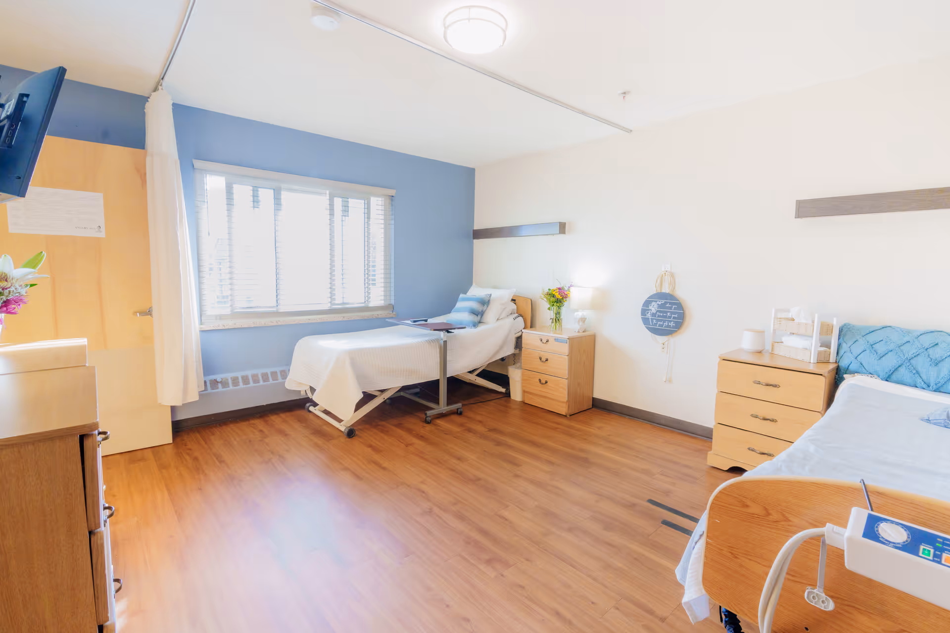 Bright, accessible care facility bedroom with two adjustable beds, wooden dressers, a window, and bedside medical equipment.