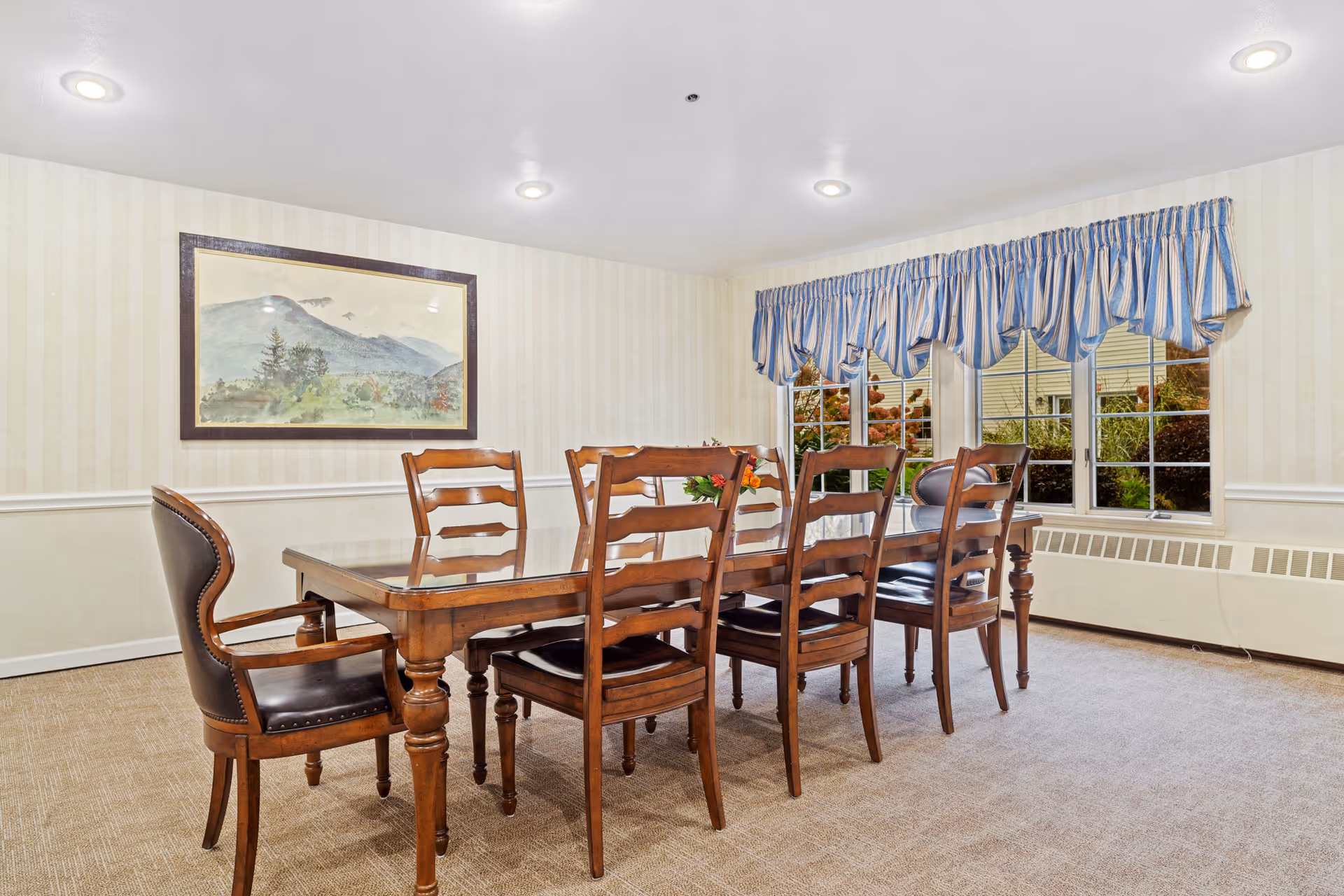 A bright dining room with a wooden table and six chairs beneath recessed ceiling lights and a window with a blue-striped valance.