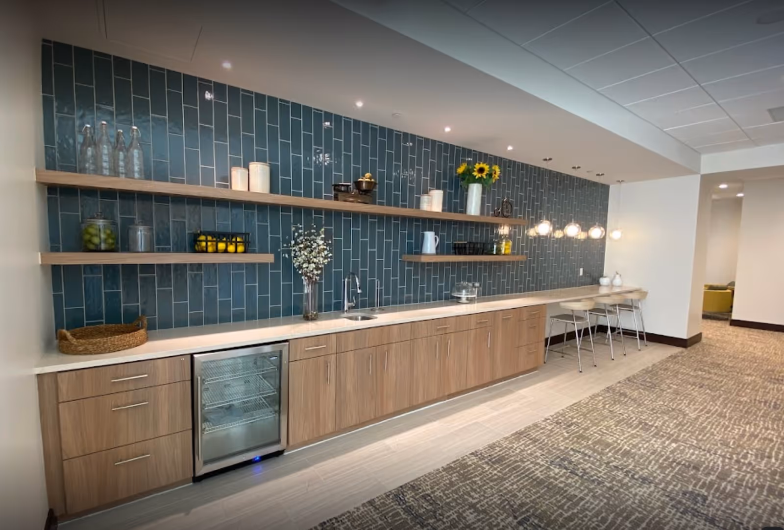 Modern communal kitchenette with wood cabinets, a blue tile backsplash, floating shelves, a sink, mini fridge and bar stools under pendant lights.