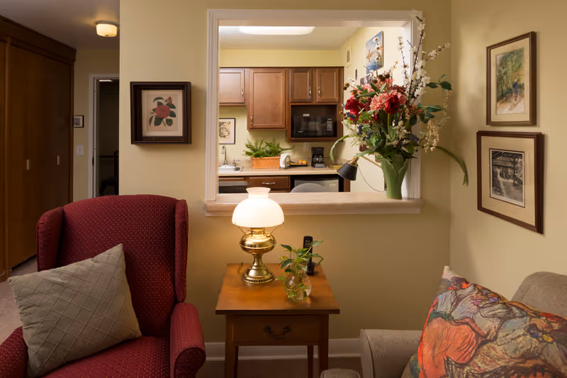 Cozy living room area with a red armchair and a gray sofa adorned with colorful pillows. A wooden side table between the chairs holds a brass lamp with a white shade and a small plant. A pass-through window opens to a kitchen with wooden cabinets, a microwave, and a coffee maker. The walls are decorated with framed artwork and a large floral arrangement sits on the kitchen counter.