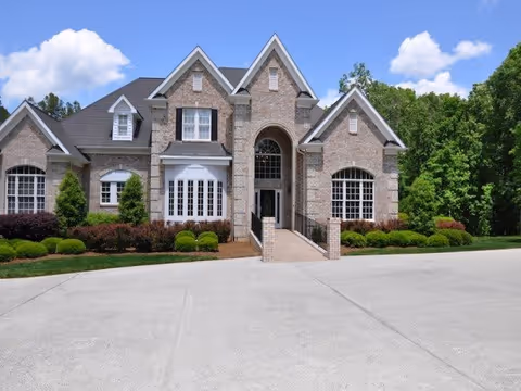 Front exterior view of a large brick building with multiple gables, large windows, and a central arched entrance. The building is surrounded by neatly trimmed bushes and trees, with a wide concrete driveway in front under a blue sky with some clouds.