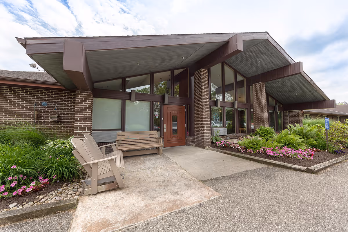Exterior view of Cedar Hill Healthcare & Rehabilitation building entrance with a brown brick facade, large windows, a covered walkway, wooden bench and chair, and landscaped flower beds with pink flowers and green plants.