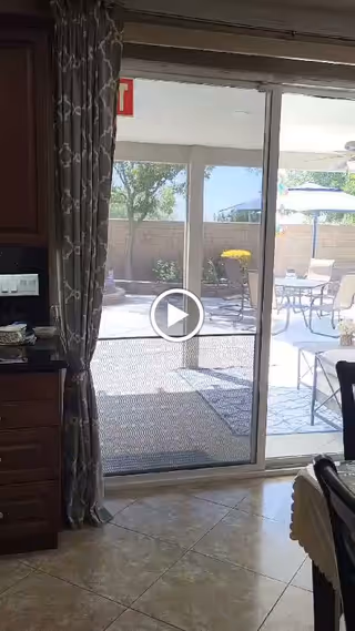 View through a sliding glass door from an interior kitchen area to an outdoor patio with a table, chairs, umbrella, and some plants along a brick wall.