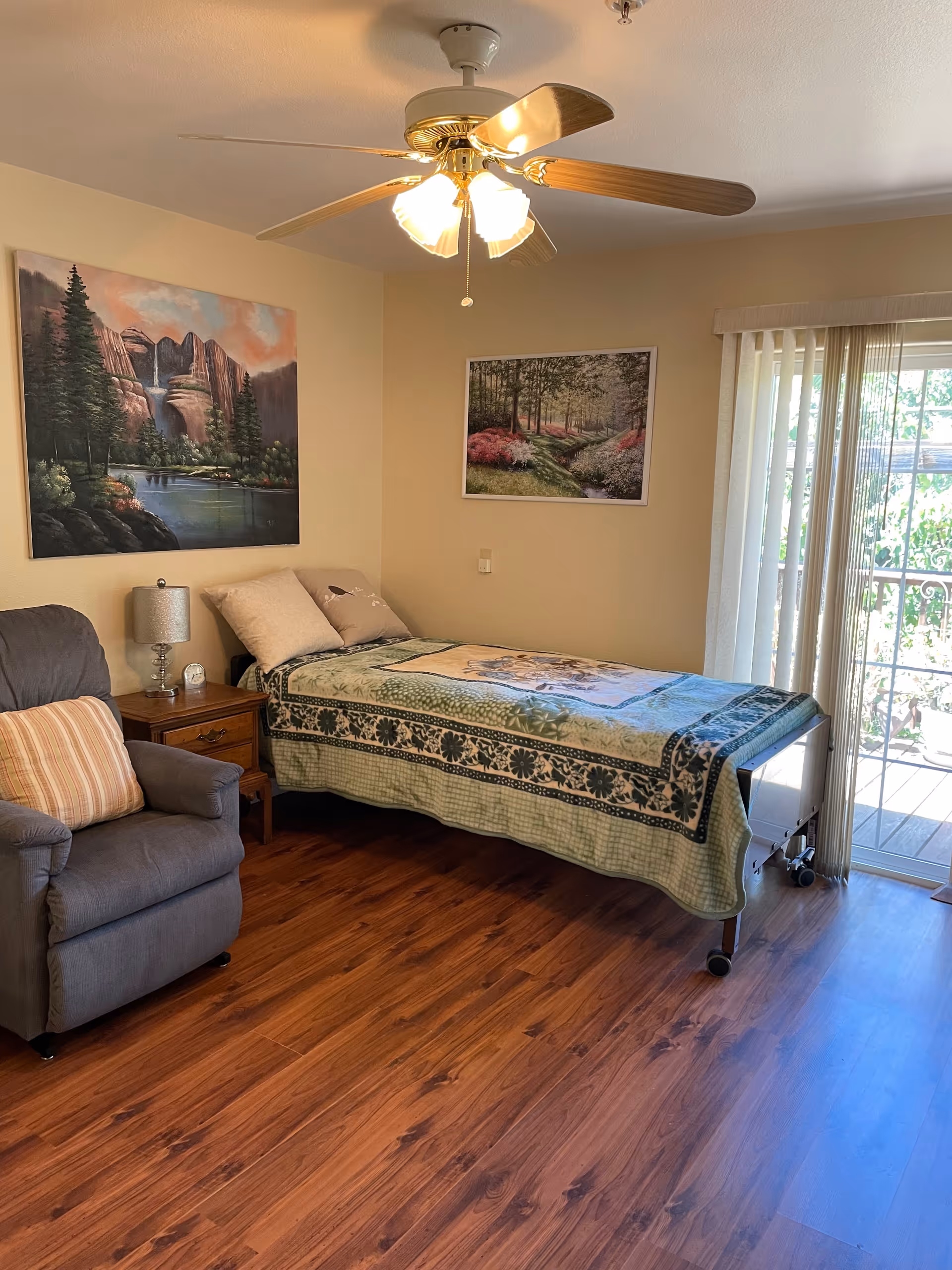A cozy bedroom with a single bed covered with a green patterned quilt, two pillows, a wooden nightstand with a lamp and clock, a gray armchair with a striped cushion, two landscape paintings on the walls, a ceiling fan with lights, and a sliding glass door with vertical blinds leading to an outdoor area.
