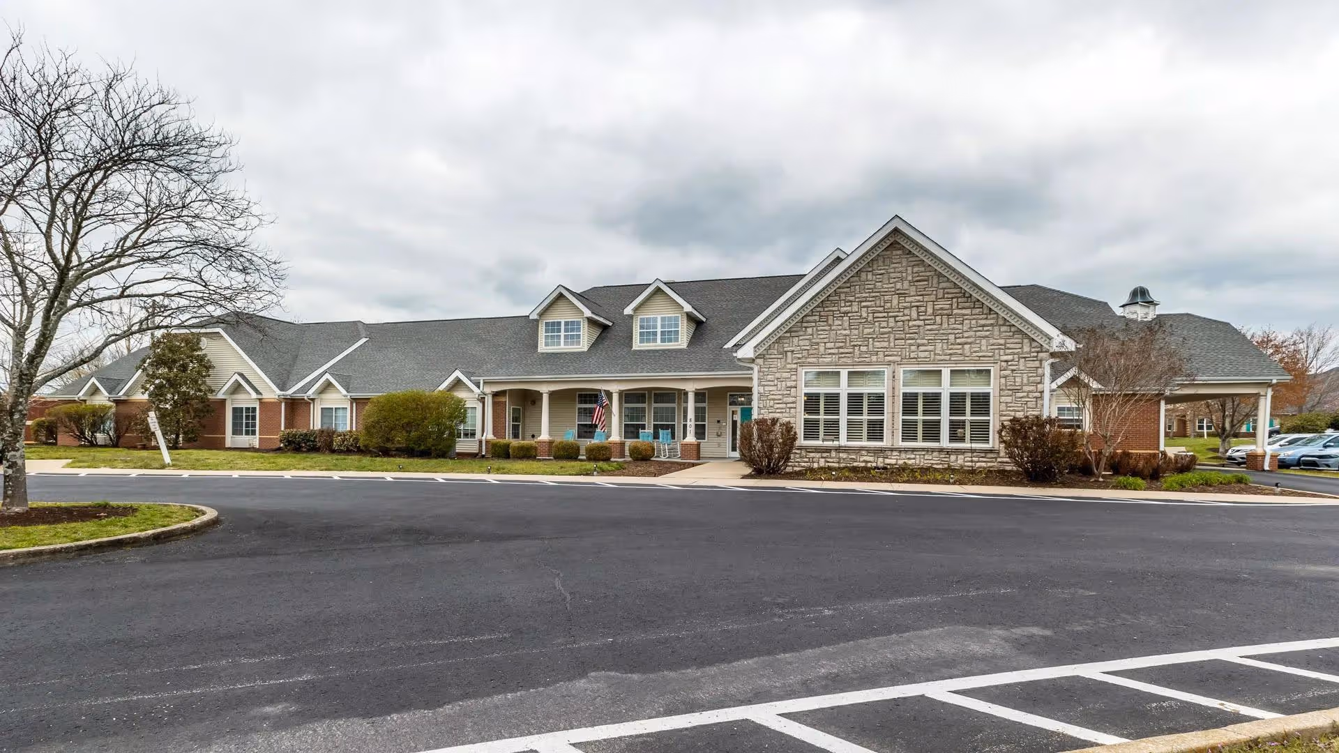 Single-story senior living facility building with a covered entrance, large windows, landscaped shrubs, and an empty parking lot under a cloudy sky.