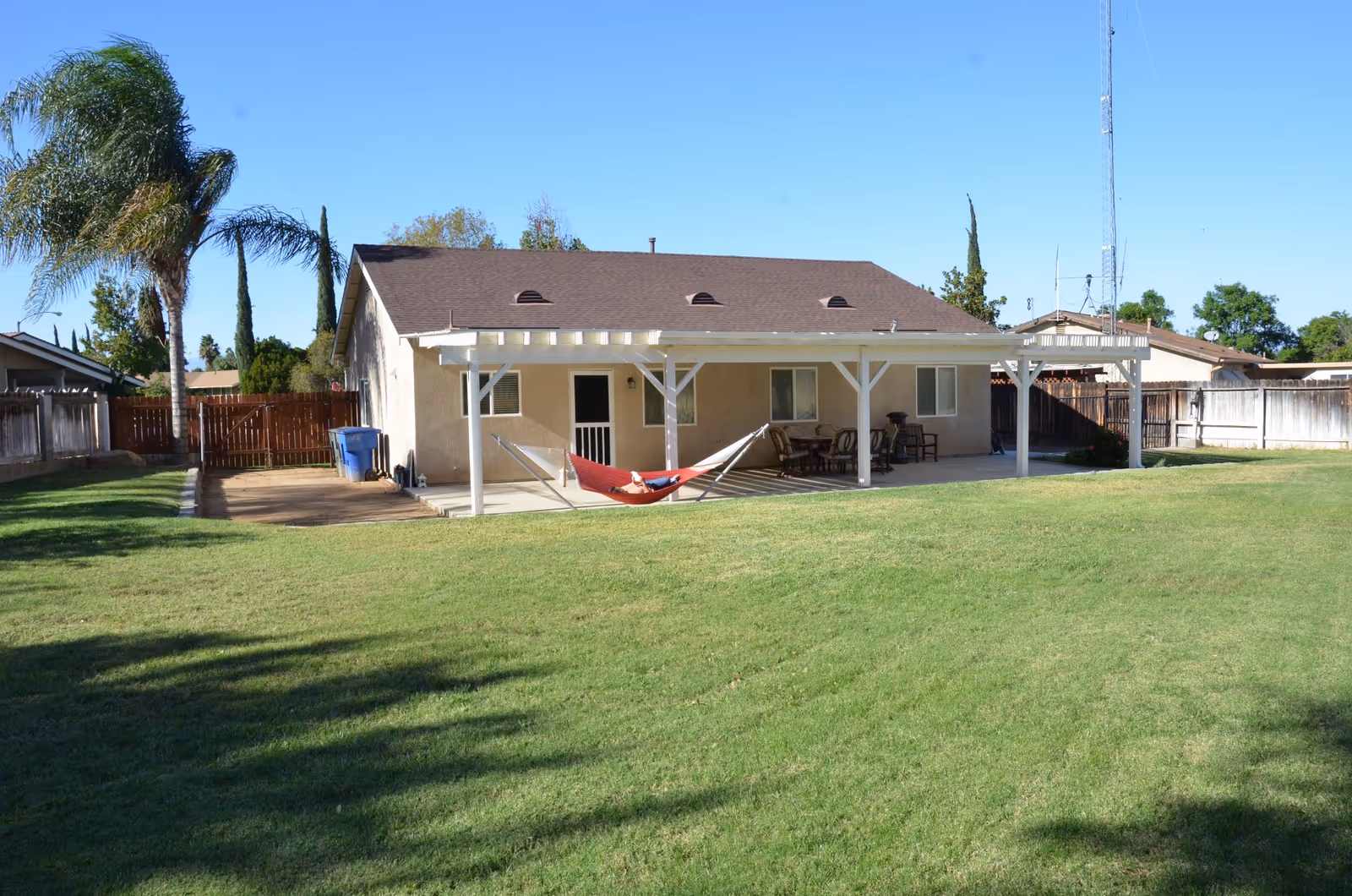 Backyard of a single-story house featuring a covered patio with a hammock and patio furniture and a large grassy lawn.
