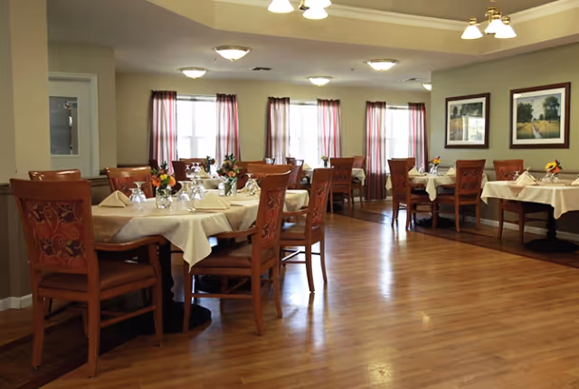 Dining room with round and rectangular tables set with white tablecloths, wooden chairs, floral centerpieces, and windows with curtains.