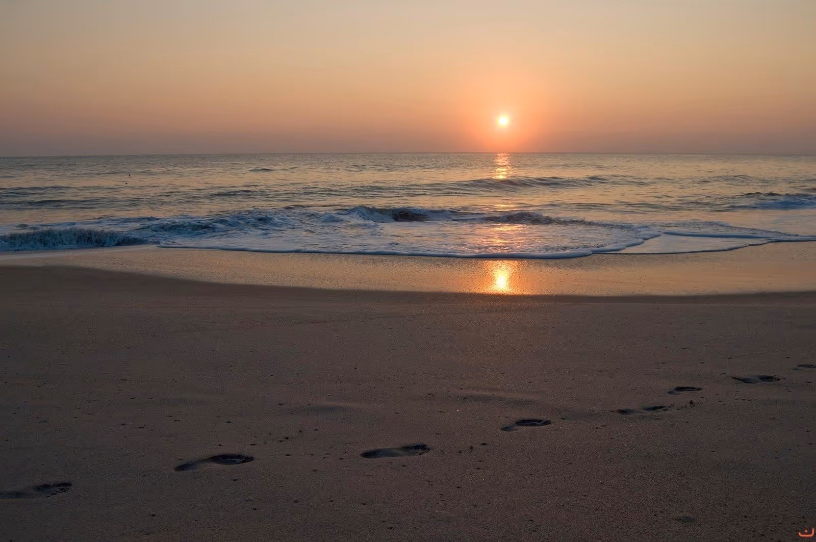 Sunset over a calm ocean with waves lapping a sandy beach and footprints in the sand.