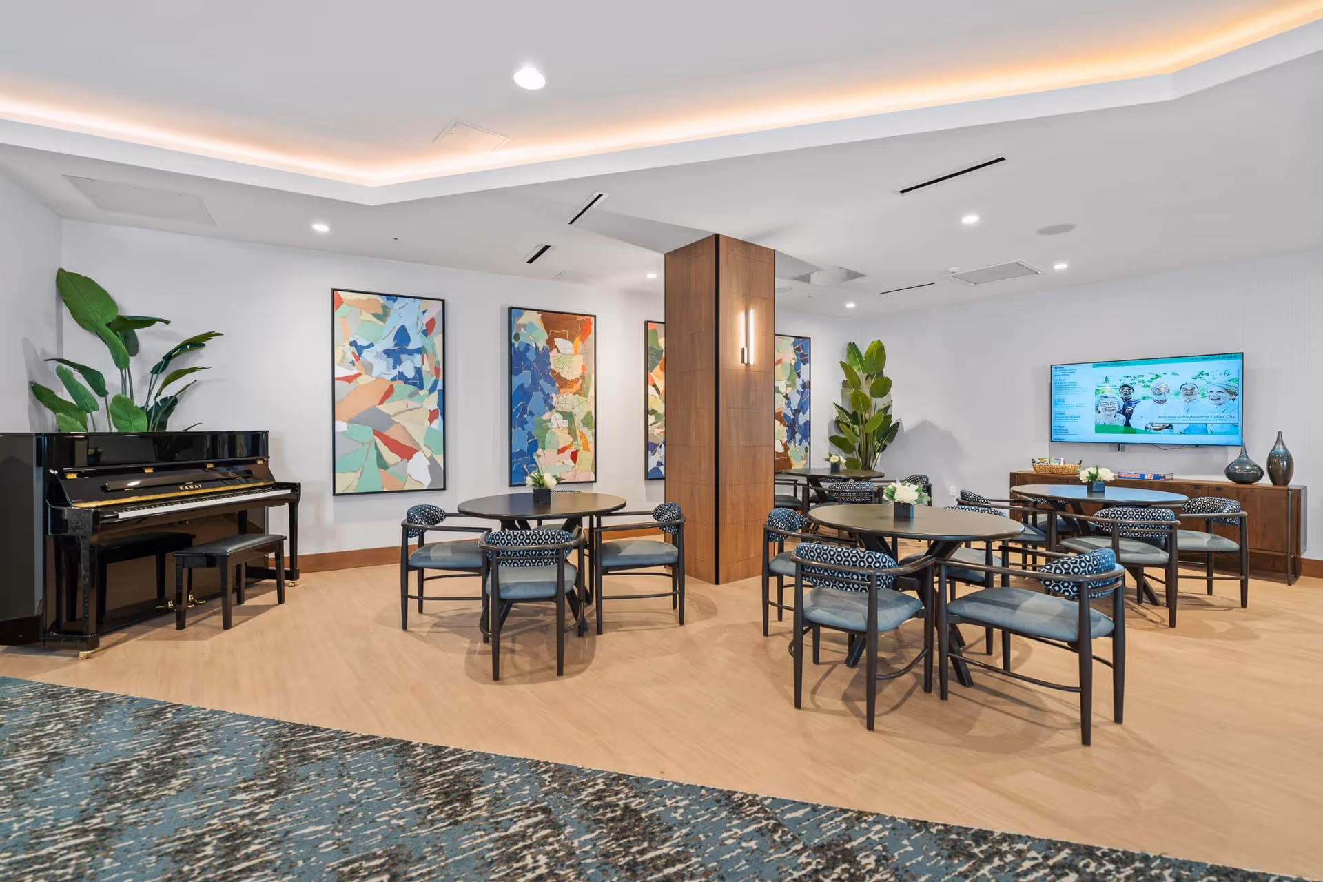 A modern common area with round tables and chairs arranged for seating. There is a black piano with a bench on the left side, large green plants, and colorful abstract artwork on the walls. A flat-screen TV is mounted on the right wall above a wooden cabinet with decorative vases and a basket.
