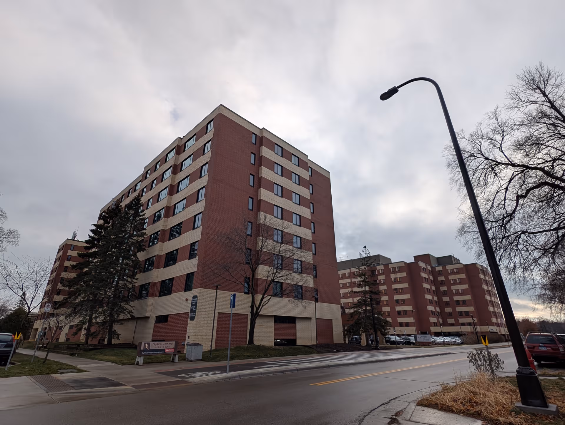 Exterior view of a multi-story brick and beige senior living facility building on a cloudy day, with a street and sidewalk in the foreground, some trees without leaves, and a streetlamp on the right side.
