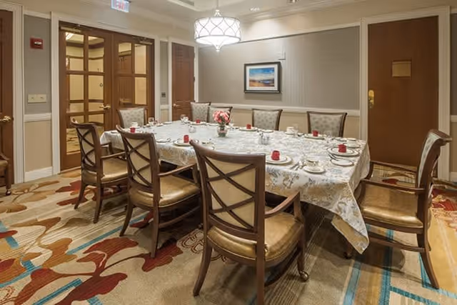 A dining room with a long rectangular table covered with a white patterned tablecloth. The table is set with plates, cups, and red napkins, and a vase with pink flowers is in the center. There are eight wooden chairs with cushioned seats around the table. The room has beige walls, a framed picture on the wall, a decorative ceiling light fixture, and a patterned carpet with various colors.