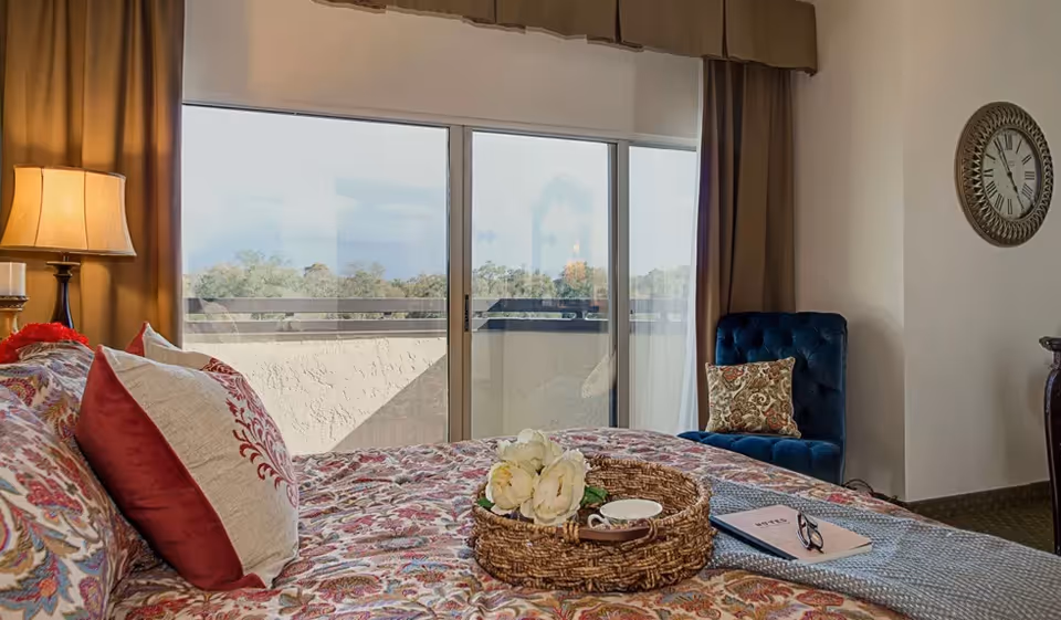 A cozy bedroom with a bed covered in a colorful patterned bedspread and multiple pillows. A wicker tray with white flowers, a cup, and a book is placed on the bed. To the right, there is a blue upholstered chair with a decorative pillow. A large sliding glass door with beige curtains leads to an outdoor balcony, and a round wall clock is mounted on the wall.