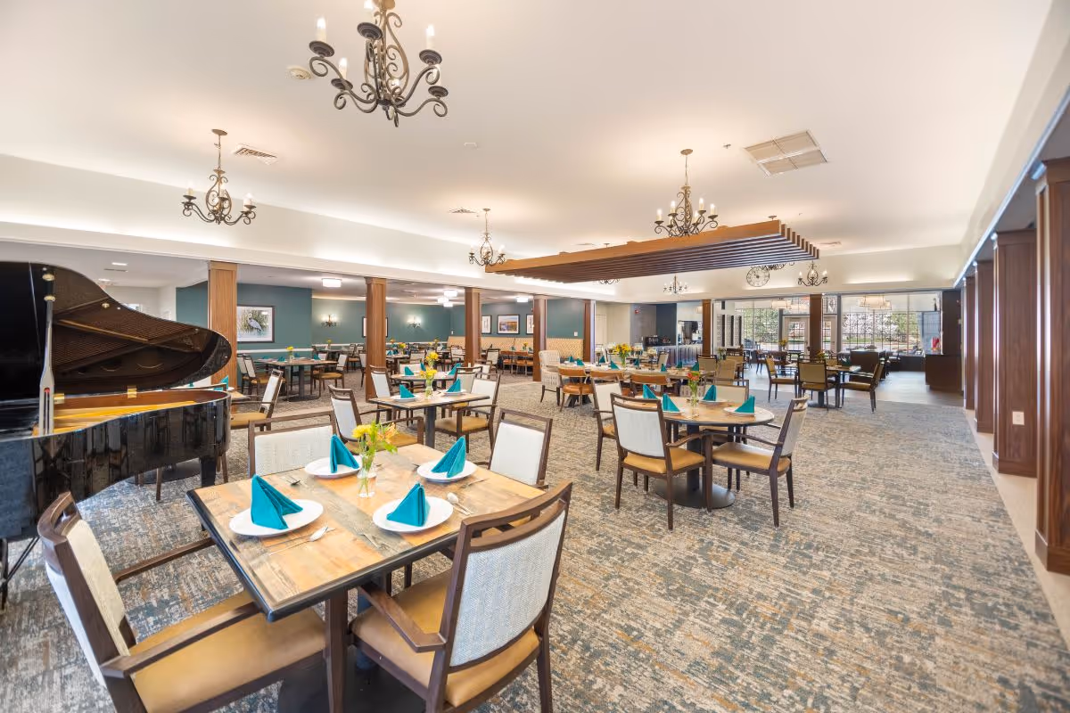 A spacious dining room in a senior living facility with multiple wooden tables set with white plates and teal folded napkins. The room features elegant chandeliers, a grand piano on the left side, and large windows allowing natural light to fill the space. The carpet has a patterned design, and there are comfortable chairs around each table. The walls are painted in a soft green color with framed artwork and sconces.