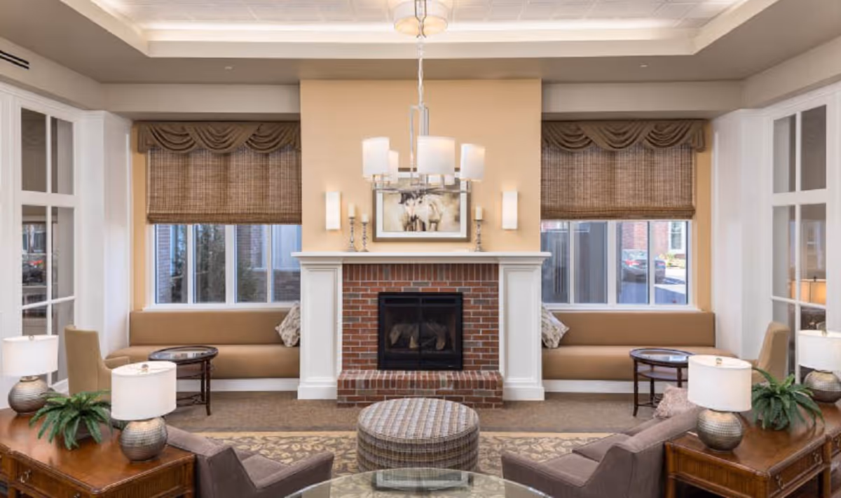 A cozy living room area with a central brick fireplace flanked by two large windows with woven shades. There are built-in bench seats under each window with cushions, two armchairs facing the fireplace, a round ottoman in the center, and side tables with lamps and plants. The room has a warm, inviting atmosphere with neutral tones and soft lighting.