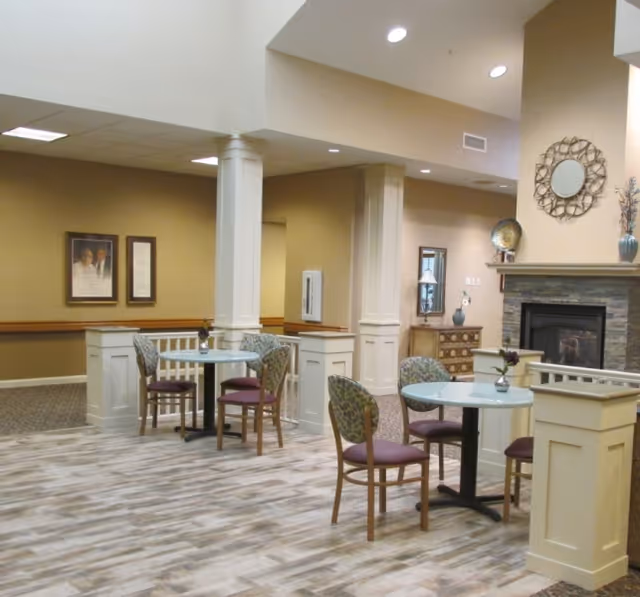 Communal dining area with round tables and chairs, a decorative fireplace, columns and wood-look flooring.
