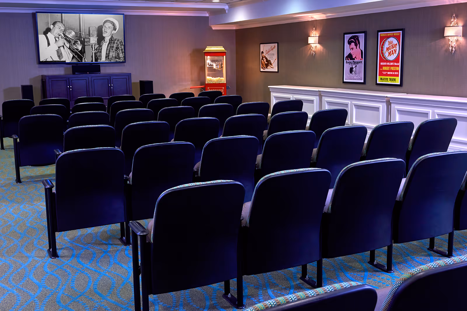 A small movie theater room with several rows of dark blue cushioned chairs facing a large screen displaying a black and white image of two musicians. There is a popcorn machine in the corner and framed vintage posters on the wall.