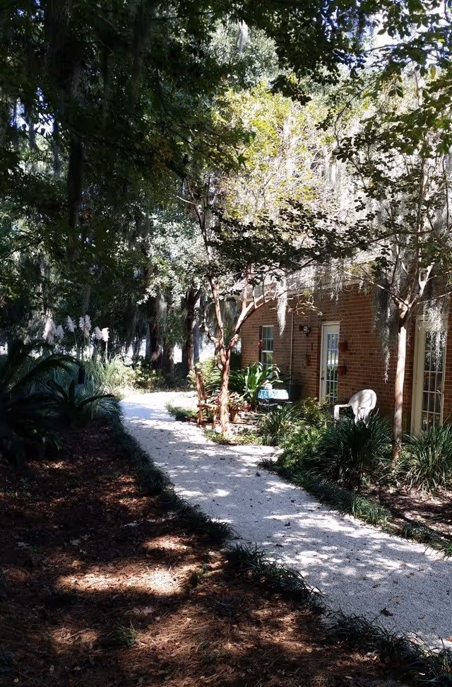 A shaded outdoor garden path lined with trees and plants, leading past a brick building with glass doors and windows. There are chairs and a small table along the path, surrounded by greenery.
