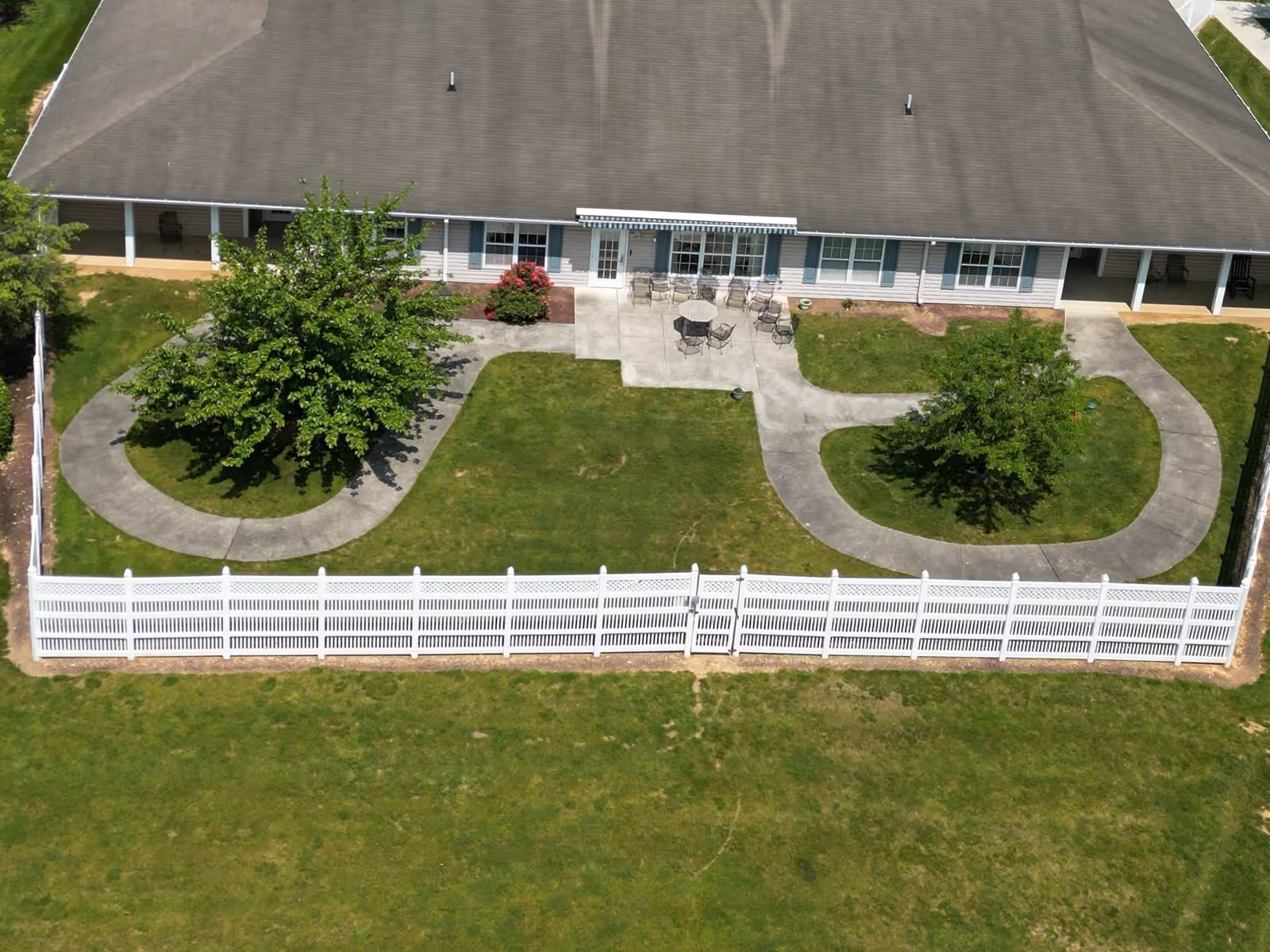 Aerial view of the backyard area of a senior living facility with a large building, a concrete patio with outdoor tables and chairs, two trees surrounded by curved concrete walkways, and a white picket fence enclosing the grassy yard.