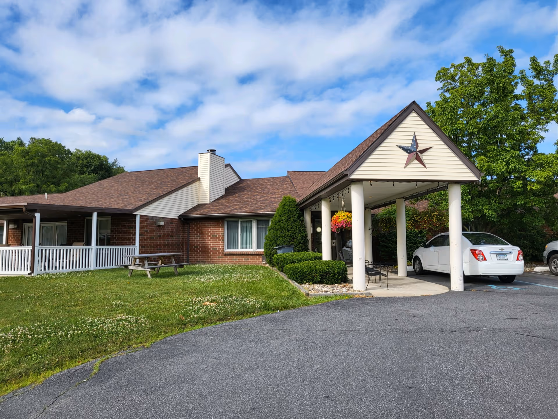 Exterior view of a single-story brick building with a covered entrance supported by four white columns. There is a decorative star on the front gable above the entrance. A white car is parked under the covered entrance, and there is a picnic table on the grassy area to the left. Trees and a partly cloudy sky are visible in the background.