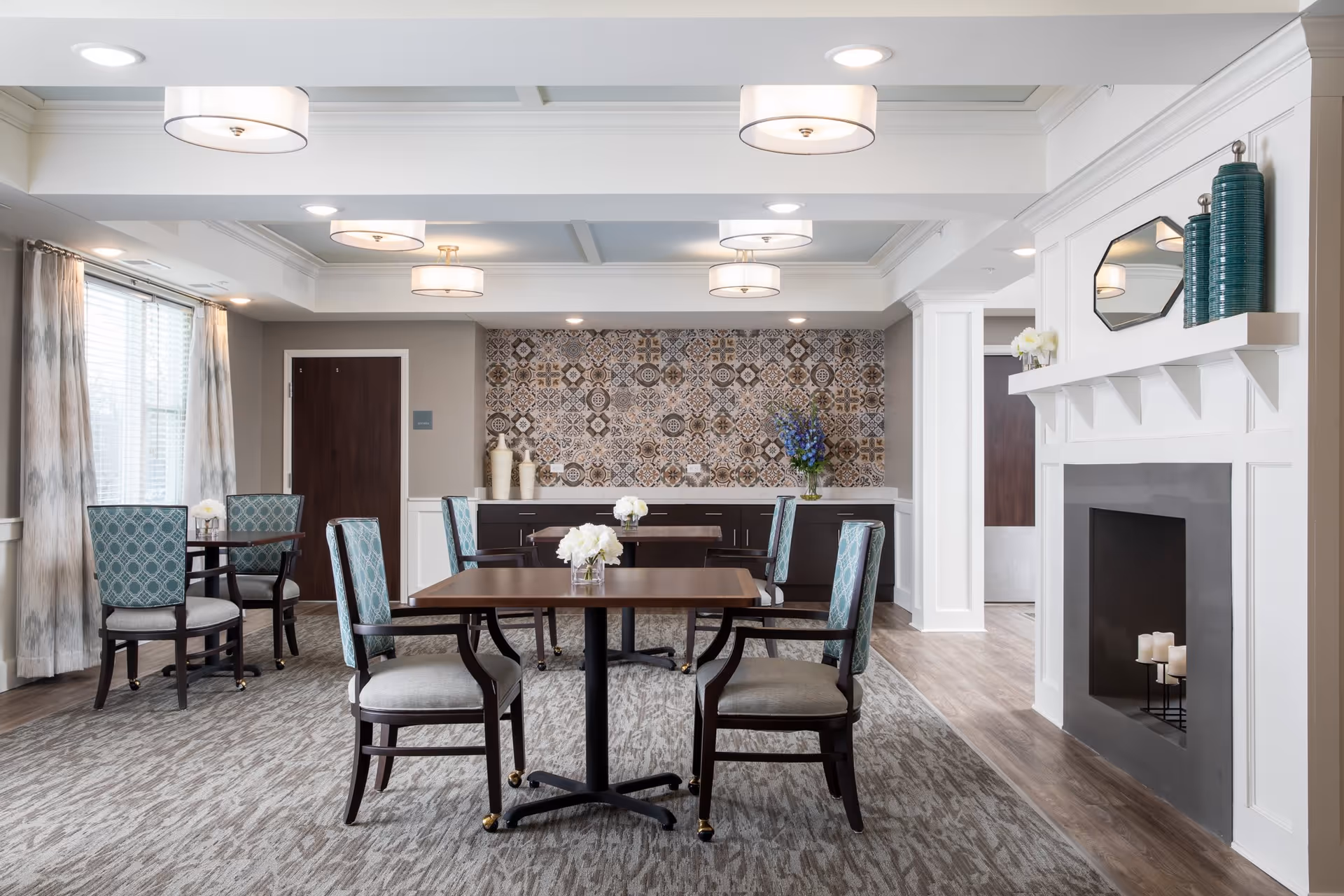A bright and elegant dining area with several wooden tables and cushioned chairs featuring blue patterned backs. The room has a decorative tiled wall, a white fireplace with candles, and vases with flowers on the tables and countertop. Large windows with curtains allow natural light to fill the space.