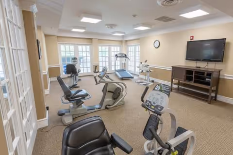 A well-lit exercise room with several cardio machines including stationary bikes, a treadmill, and an elliptical. The room has beige walls, carpeted floor, large windows letting in natural light, a wall-mounted clock, and a flat-screen TV on a wooden stand.