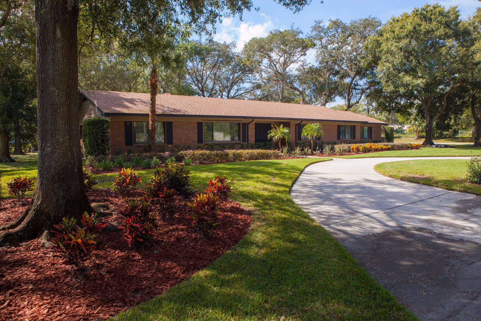 Single-story brick building with a brown shingled roof surrounded by well-maintained landscaping including green grass, small palm trees, and red-leafed plants. A curved concrete driveway leads up to the entrance of the building under a partly cloudy blue sky.