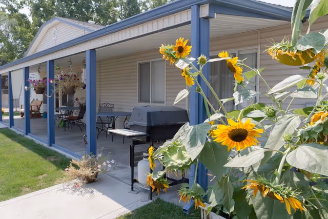 Covered patio with seating, a grill, and hanging planters in front of a single-story building, with sunflowers in the foreground.