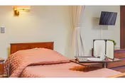 A neatly made bed with a pink quilted bedspread in a room. There is a wooden headboard, a wall-mounted light fixture above the bed, and a window with beige curtains. A flat-screen TV is mounted on the wall, and a wooden dresser with a tray holding a menu or informational booklet is visible.