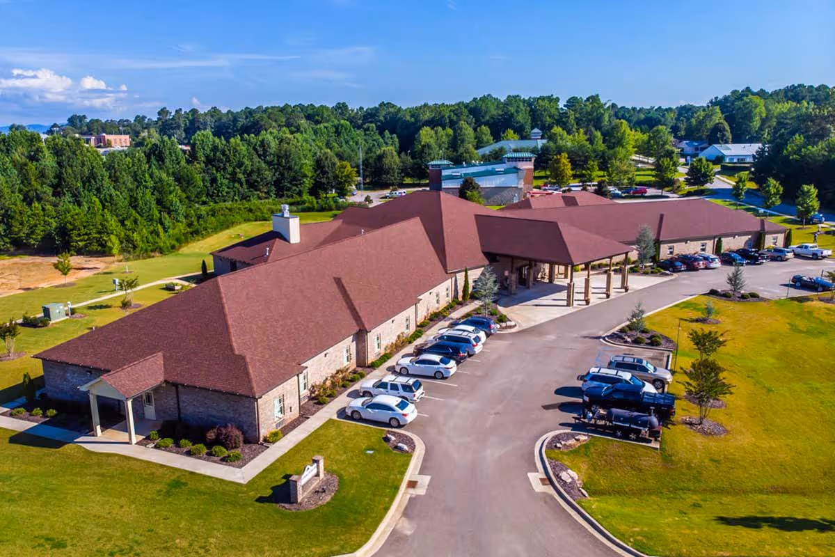 Aerial view of a large single-story assisted living facility with a red roof, surrounded by green lawns and trees. Several cars are parked in the parking lot in front of the building under a clear blue sky.