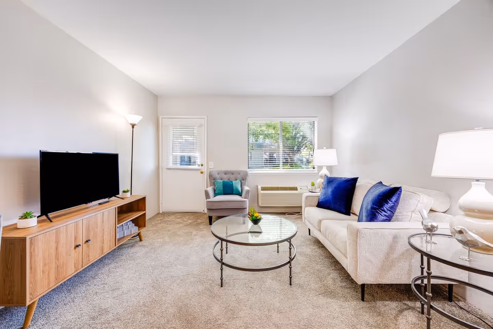 A bright living room with beige carpet and walls, featuring a light beige sofa with two dark blue pillows, a glass coffee table, a gray armchair with a teal pillow, a wooden TV stand with a flat-screen TV, and two lamps providing warm light. A window with blinds and a door are visible in the background.