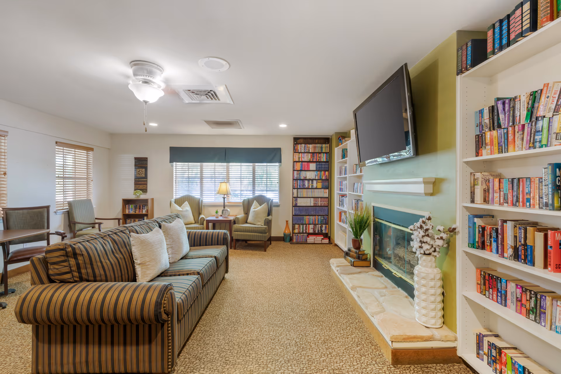 Cozy senior living common room with a striped sofa, armchairs by a window, bookshelves, a fireplace and a wall-mounted TV.