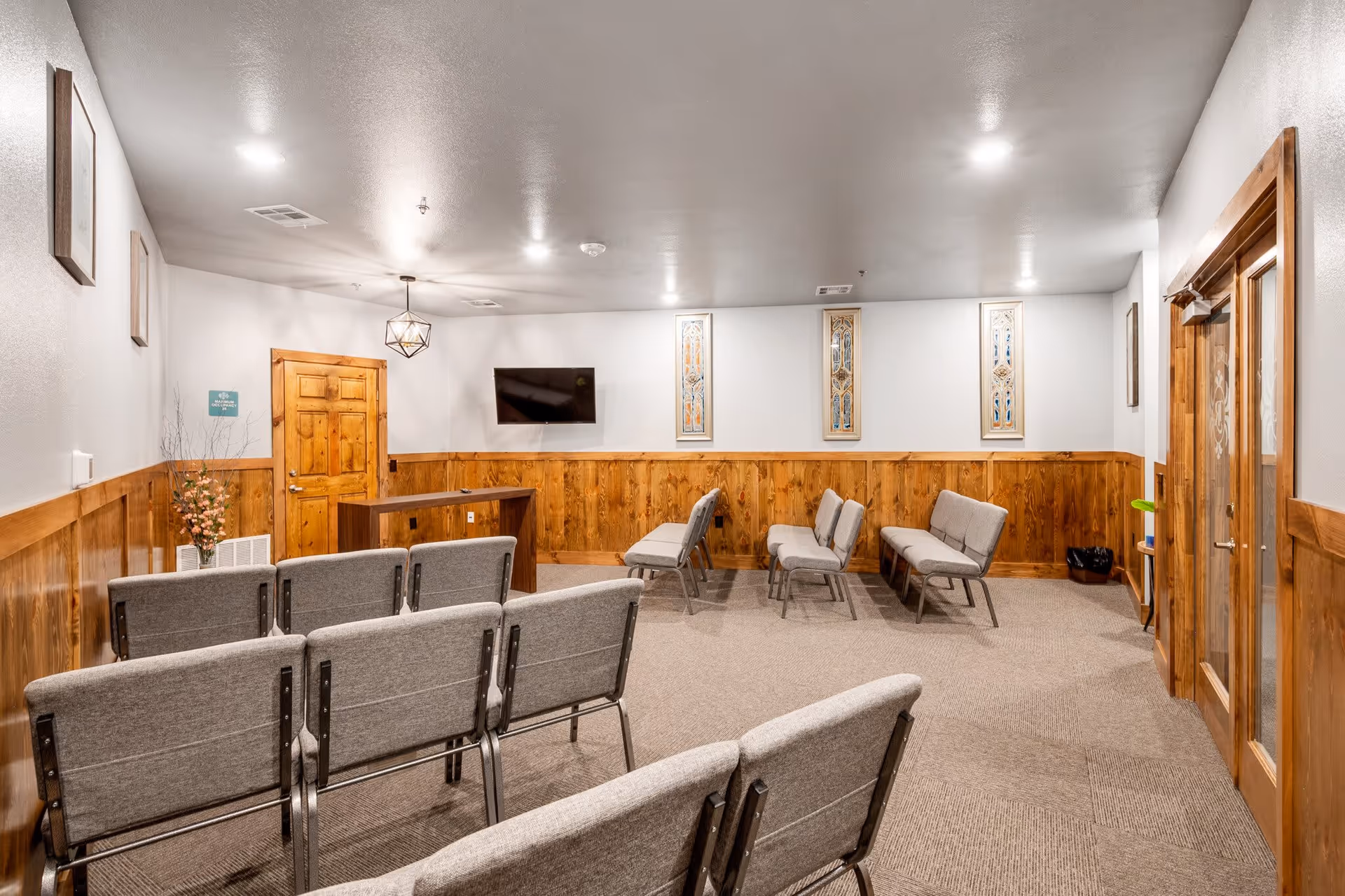 A small meeting or activity room with rows of gray cushioned chairs facing a wooden podium and a wall-mounted TV. The room has wood paneling on the lower half of the walls, beige carpet, and decorative wall art. There is a wooden door on the left and glass double doors on the right.