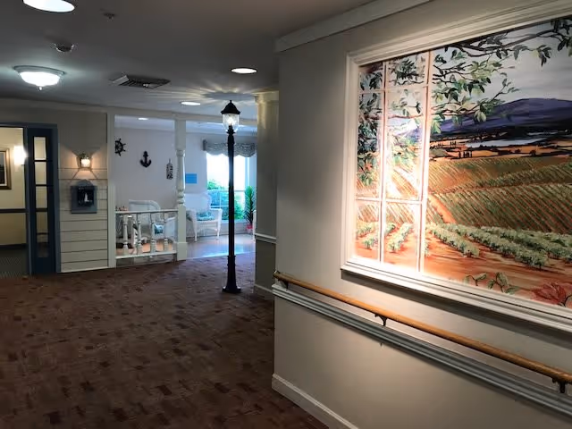 Interior hallway of a senior living facility with brown carpet and white walls. On the right wall, there is a large framed painting depicting a scenic landscape with fields and mountains. The hallway features a wooden handrail along the wall. At the end of the hallway, there is a small seating area with white chairs and nautical-themed decorations such as an anchor and ship wheel on the wall. A black streetlamp-style light fixture stands near the seating area.