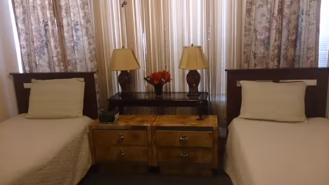 A bedroom with two single beds separated by two wooden nightstands. Each bed has a white pillow and beige bedspread. Behind the nightstands is a small table with two matching lamps and a vase with red flowers. The background features windows covered with floral curtains and vertical blinds.