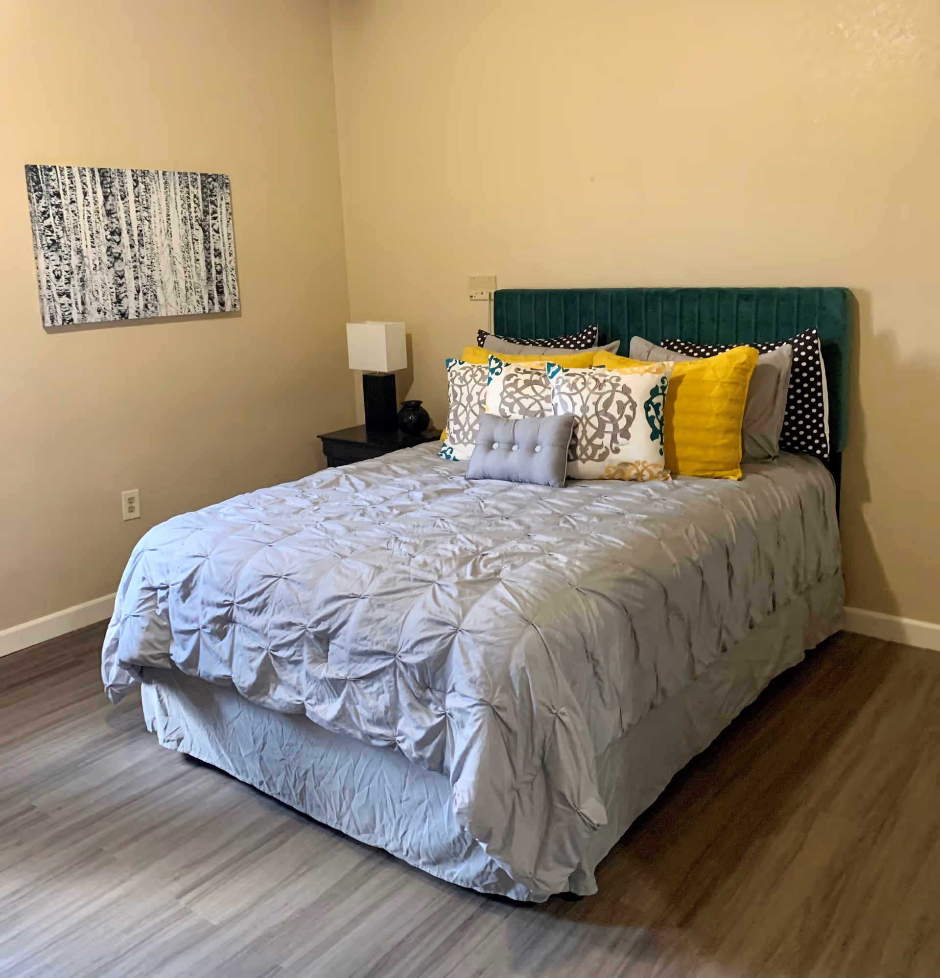 A neatly made bed with a green upholstered headboard, gray comforter, and multiple decorative pillows in yellow, gray, white, and patterned designs. Next to the bed is a black nightstand with a white square lamp and a small black vase. A black and white abstract painting hangs on the beige wall above the nightstand. The room has wood flooring and beige walls.