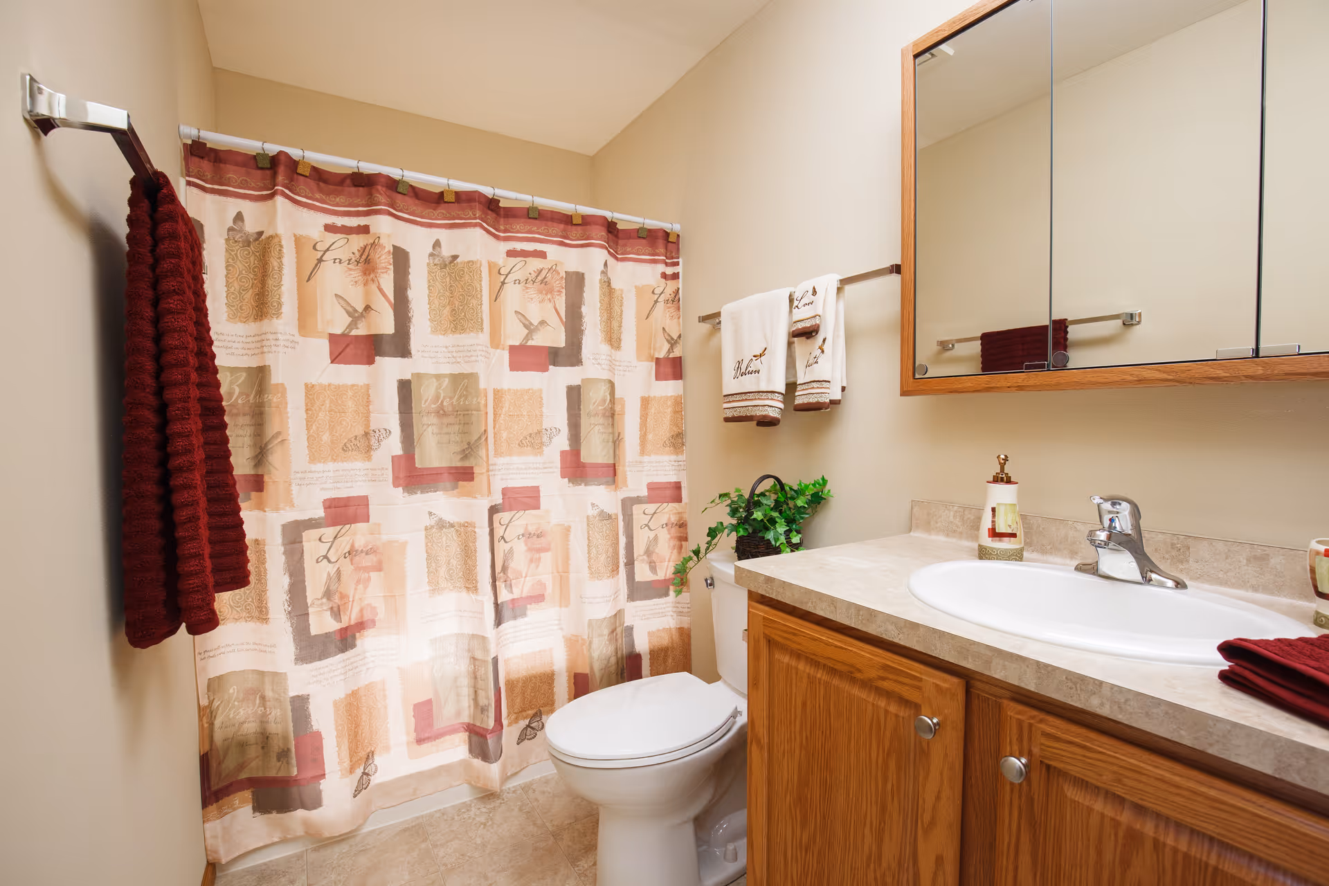 A clean bathroom featuring a toilet, a sink with a countertop and wooden cabinets, a mirror above the sink, a shower with a patterned curtain, and several towels hanging on racks. There is also a small green plant placed on the toilet tank.